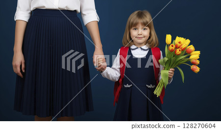 Young schoolgirl in uniform with flowers and backpack holding her mother's hand. First day at school. 128270604