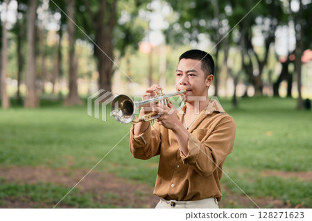 Young man playing trumpet outdoors in a peaceful green park 128271623