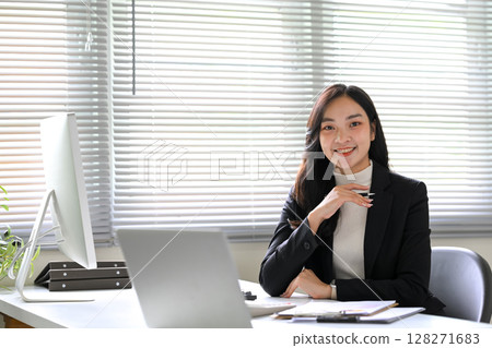 Confident businesswoman in formal attire smiling at her desk in a bright modern office 128271683
