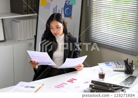 Focused young businesswoman analyzing charts and documents at her desk 128271684