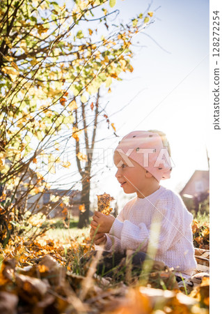 Adorable preschooler girl enjoying nice and sunny autumn day outdoors. Happy child sitting on the ground covered with autumn leaves. Outdoor fall activities for kids 128272254