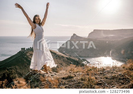 A woman in a white dress stands on a rocky hill overlooking the ocean. She is smiling and she is happy. 128273496