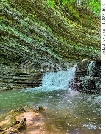 Waterfall flows over textured rock layers in peaceful forest. Surrounding greenery adds vibrancy to serene scene. Concept of nature exploration, outdoor adventure, eco-tourism. 128273698