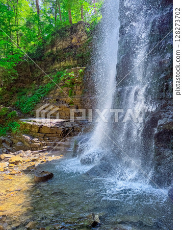 Water flows down rocky cliff creating waterfall in vibrant natural setting. Sunlight filters through trees casting shadows on clear water pool. Concept of eco-tourism, outdoor adventure, relaxation. Water flows down rocky cliff creating waterfall in vibrant natural setting. Sunlight filters through trees casting shadows on clear water pool. Concept of eco-tourism, outdoor adventure, relaxation. 128273702