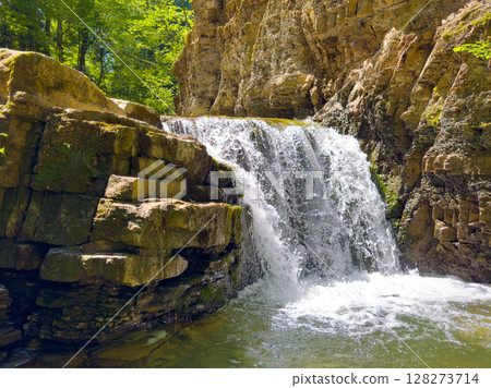 Waterfall flows down rocky surface into clear pool surrounded by vibrant green foliage. Peaceful forest atmosphere invites relaxation and adventure. 128273714