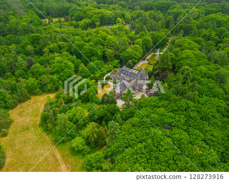 An aerial view of a historic mansion surrounded by lush green trees and a beautifully landscaped garden with a fountain, walking paths, and parking area. The scene shows vibrant summer greenery. 128273916