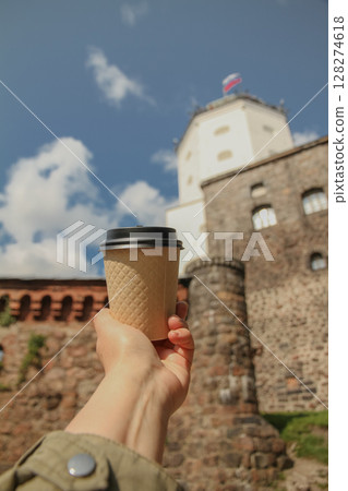 Brown paper cup of natural coffee in women's hand on against backdrop of Olaf's Tower in city Vyborg in blur, Russia on sunny day Brown paper cup of natural coffee in women's hand on against backdrop of Olaf's Tower in city Vyborg in blur, Russia on sunny day 128274618