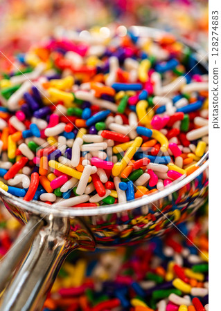 Clear glass scoop resting in a pile of rainbow sprinkles, focused on the texture and vivid colors. 128274883