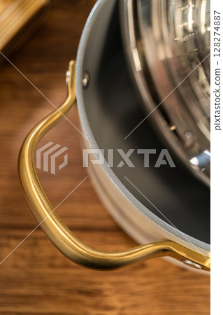 Top side view of a ceramic pot with a golden handle and a stainless steel steamer insert. The setup is styled on a wood surface with soft light. Top side view of a ceramic pot with a golden handle and a stainless steel steamer insert. The setup is styled on a wood surface with soft light. 128274887
