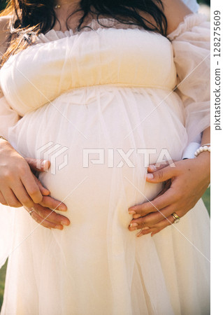 Expecting mother gently cradles her baby bump in a peaceful outdoor setting during golden hour 128275609