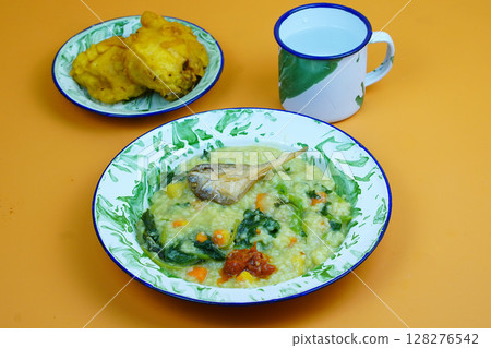 A traditional Indonesian meal featuring Bubur Manado or porridge with vegetables and salted fish on a classic plate, accompanied by fried tofu and a glass of water. 128276542
