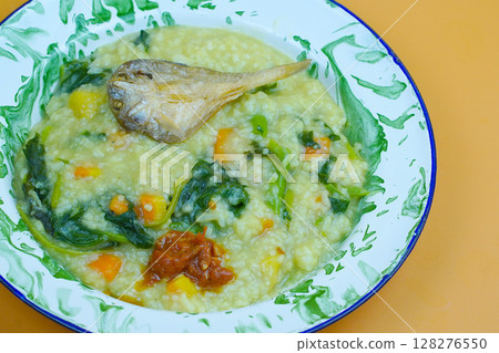 A traditional Indonesian meal featuring Bubur Manado or porridge with vegetables and salted fish on a classic plate, accompanied by fried tofu and a glass of water. A traditional Indonesian meal featuring Bubur Manado or porridge with vegetables and salted fish on a classic plate, accompanied by fried tofu and a glass of water. 128276550