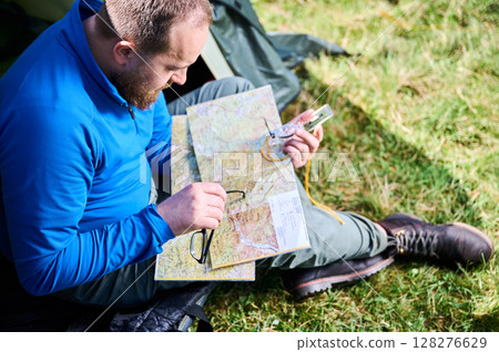 Close up man in glasses closely examines map while holding compass. He sits by his tent, deeply focused on navigation. Lush greenery, suggesting serene outdoor adventure. Close up man in glasses closely examines map while holding compass. He sits by his tent, deeply focused on navigation. Lush greenery, suggesting serene outdoor adventure. 128276629