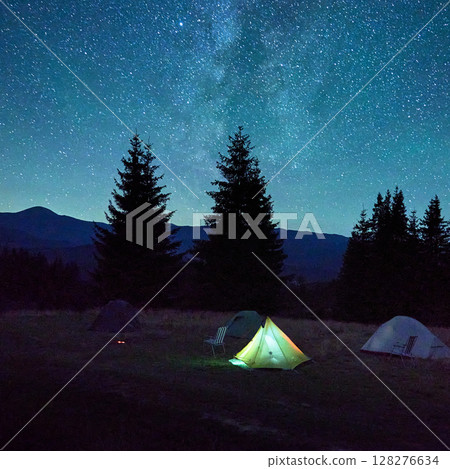 Few tents on grassy clearing under dazzling, starry sky with Milky Way clearly visible. Tall pine trees and distant mountains frame scene, creating tranquil and awe-inspiring night in nature. 128276634