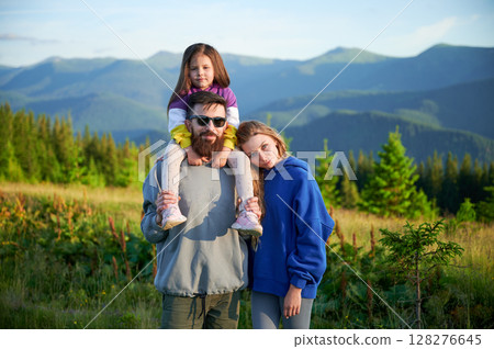 Family stands together in scenic meadow with backdrop of rolling mountains. Father carries his daughter on shoulders, both smiling and enjoying view, while mother stands beside them. Family stands together in scenic meadow with backdrop of rolling mountains. Father carries his daughter on shoulders, both smiling and enjoying view, while mother stands beside them. 128276645