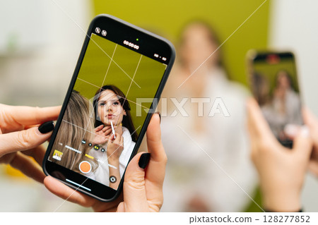 Female hands holding smartphone recording video of beautician applying makeup on model face, creating content for social media or online tutorial in cosmetology clinic. Concept of beauty treatment. 128277852