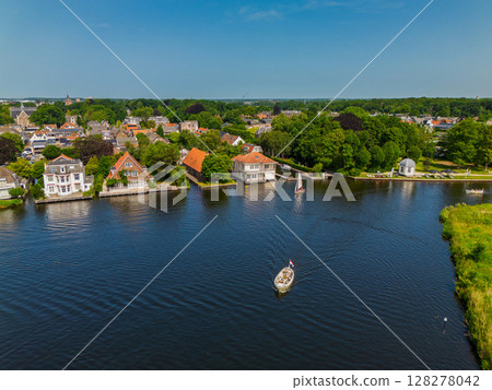 A yacht with a white sail sails along the canal. A beautiful windmill. Aerial view of narrow canal running through lush green fields and a small forest leading to peaceful Dutch village with a marina 128278042