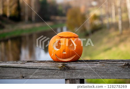 Smiling carved Halloween pumpkin on wooden railing by the river Smiling carved Halloween pumpkin on wooden railing by the river 128278269