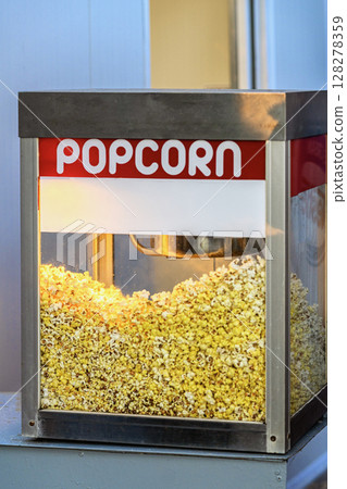 Popcorn Machine Close-Up. Popcorn machine full of golden popped kernels glows warmly behind transparent glass. Concept of snack, retro style, comfort food joy Popcorn Machine Close-Up. Popcorn machine full of golden popped kernels glows warmly behind transparent glass. Concept of snack, retro style, comfort food joy 128278359