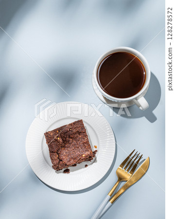 Chocolate brownie on a plate with fork on a blue background with cup of coffee, cutlery and shadows. 128278572