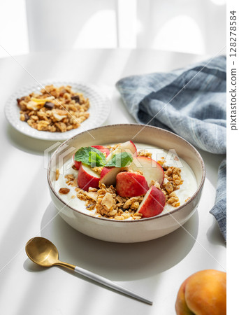 Natural greek yogurt with granola or muesli and peach in a bowl on a white table near window 128278584
