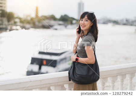 Happy solo Southeast Asian teenager woman travel blogger uses smartphone. Local cultural experience wait for express boat on Chao Phraya River in Bangkok Thailand for Asia tourism 128279242