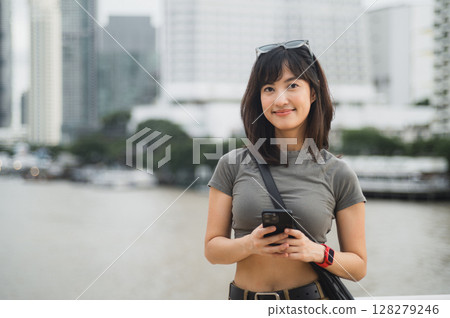 Happy young southeast asian woman teenager blogger using smartphone searching guide. World travel explorer at Chao Phraya River Bangkok asia tourism concept for local cultural experience Happy young southeast asian woman teenager blogger using smartphone searching guide. World travel explorer at Chao Phraya River Bangkok asia tourism concept for local cultural experience 128279246