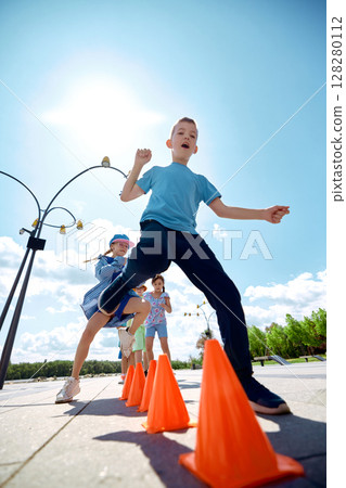 Kids engaging in outdoor activities with orange cones under the sunlit park. Kids engaging in outdoor activities with orange cones under the sunlit park. 128280112