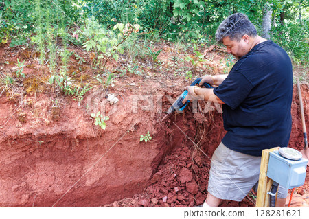 Person uses jackhammer tool to dig into red soil, revealing layer patterns roots in yard full 128281621
