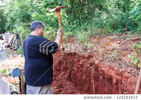 Contractor is working hard with pickaxe to remove soil roots near construction site surrounded by trees. 128281623