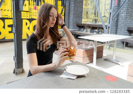 A Young Woman Happily Enjoying a Delicious and Absolutely Appetizing Burger in a Trendy Outdoor Cafe Setting 128283385