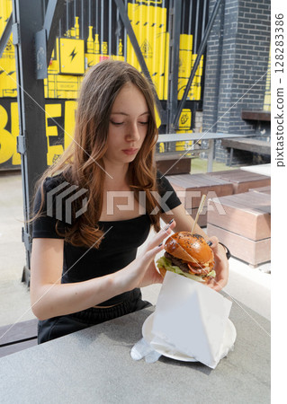 A Young Woman Happily Enjoying a Delicious and Absolutely Appetizing Burger in a Trendy Outdoor Cafe Setting A Young Woman Happily Enjoying a Delicious and Absolutely Appetizing Burger in a Trendy Outdoor Cafe Setting 128283386