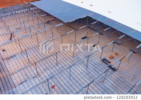 Workers assemble steel framework for warehouse under work day, with construction equipment close by. 128283912