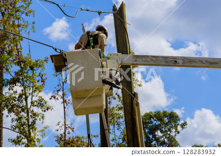 Worker in bucket truck is repairing electrical lines amid trees while sun shines brightly above. Worker in bucket truck is repairing electrical lines amid trees while sun shines brightly above. 128283932