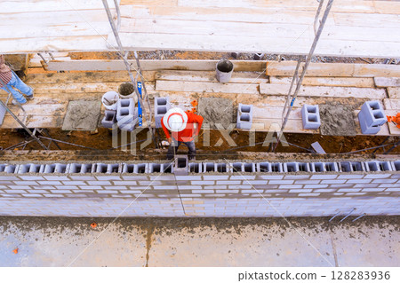 Laborers work on constructing brick wall using scaffolding, blocks, mortar in an active construction area. 128283936