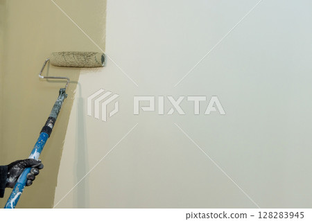 Contractor uses paint roller to apply light paint to wall while renovating room indoors. 128283945