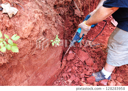 Worker uses power jackhammer to excavate red clay soil while construction landscaping techniques. Worker uses power jackhammer to excavate red clay soil while construction landscaping techniques. 128283958