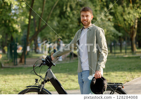Smiling man stands with electric bicycle in lush green park during daytime with trees in the background Smiling man stands with electric bicycle in lush green park during daytime with trees in the background 128284008