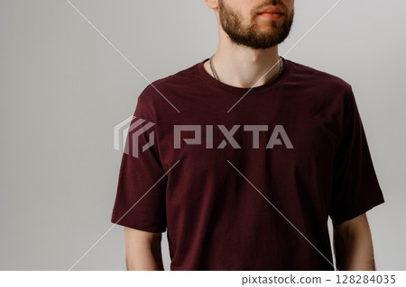 Casual male model showcasing a burgundy t-shirt against a simple gray backdrop during a studio photoshoot 128284035