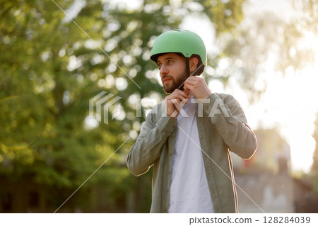 Young man adjusts helmet while preparing to ride a bike in park during golden hour 128284039