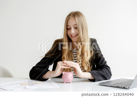 Young woman smiling while saving money in a pink piggy bank at a desk with documents and a laptop during a productive work session 128284040
