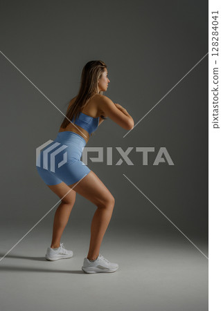 Woman performing squats in a studio setting with neutral background, showcasing fitness routine and athletic wear 128284041