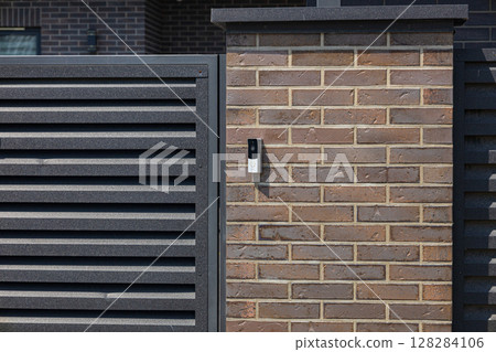 A contemporary intercom is mounted on a brick wall adjacent to a slatted gate in a well-maintained suburban area 128284106