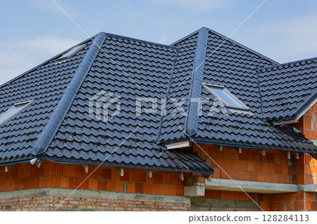 Metal roofing tiles cover a newly constructed home with multiple slopes and skylights, showcasing contemporary design elements against a blue sky 128284113