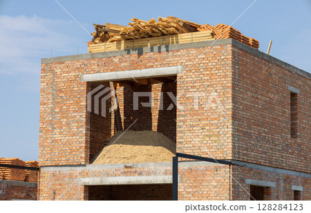Brick building is under construction, showcasing raw materials like sand and wooden planks on the rooftop against a sunny backdrop 128284123
