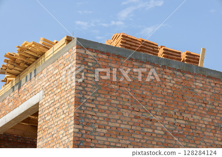 Workers are busy constructing a brick building, stacking wooden materials on the roof against a clear blue sky backdrop Workers are busy constructing a brick building, stacking wooden materials on the roof against a clear blue sky backdrop 128284124