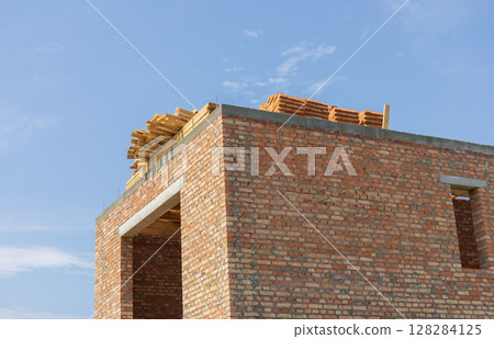 A brick structure stands incomplete with wooden beams positioned on its roof under a bright blue sky, showcasing ongoing construction work A brick structure stands incomplete with wooden beams positioned on its roof under a bright blue sky, showcasing ongoing construction work 128284125