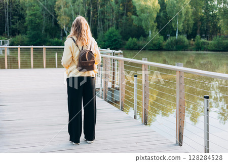 A young happy blonde woman standing on a bridge near a lake on a sunny day. Beautiful morning relax, Full body 128284528