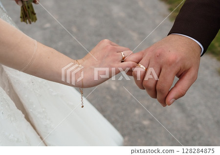 Wedding Hands Holding Intertwined with Gold Rings, White Dress, Romance, Commitment, Love, Marriage, Celebration, Outdoor Ceremony, Couple, Brown Coat 128284875