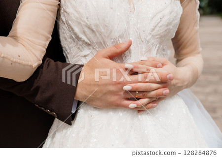 Intimate Wedding Moment: Couple Embracing with Focused Hands Holding Wedding Rings, Luxurious Dress, Brown Suit, Sparkling Texture. 128284876